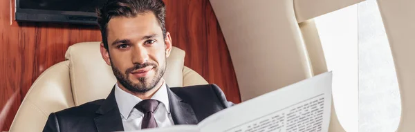 Panoramic shot of handsome businessman in suit holding newspaper in private plane — Stock Photo