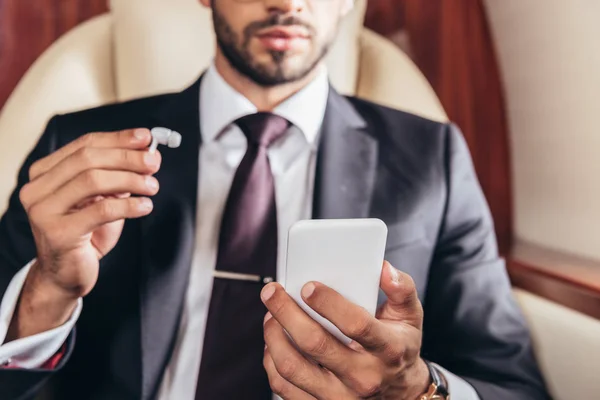 Vista recortada del hombre de negocios sosteniendo auriculares y el uso de teléfonos inteligentes en avión privado - foto de stock