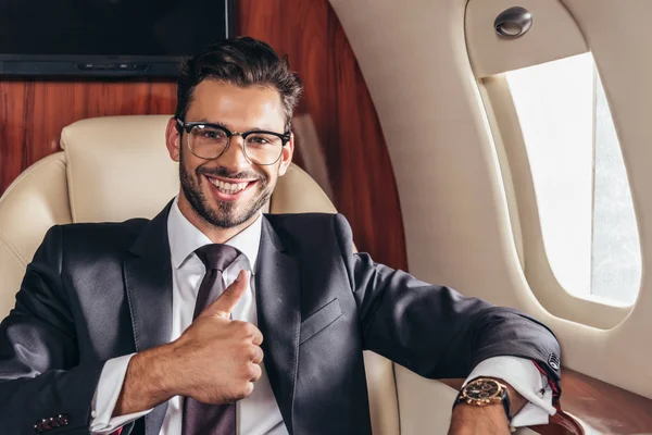 Guapo hombre de negocios en traje sonriendo y mostrando el pulgar hacia arriba en avión privado - foto de stock