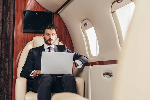Messa a fuoco selettiva di bell'uomo d'affari in giacca e cravatta utilizzando laptop in aereo privato — Foto stock