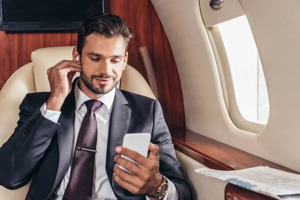 Красивый бизнесмен в костюме слушает музыку и использует смартфон в частном самолете — стоковое фото