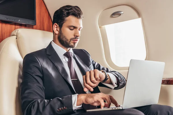 Handsome businessman in suit using laptop and looking at wristwatch in private plane — Stock Photo