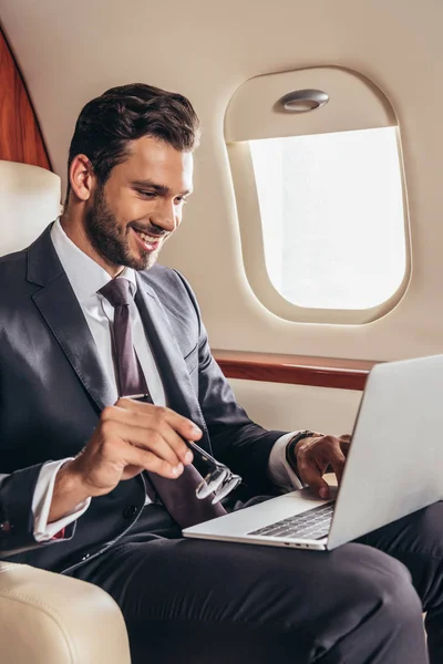 Hombre de negocios sonriente en traje usando el ordenador portátil y sosteniendo gafas en avión privado - foto de stock