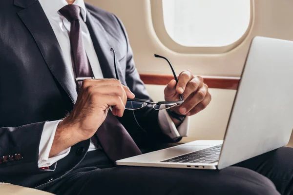 Vue recadrée de l'homme d'affaires en costume avec ordinateur portable tenant des lunettes dans un avion privé — Photo de stock
