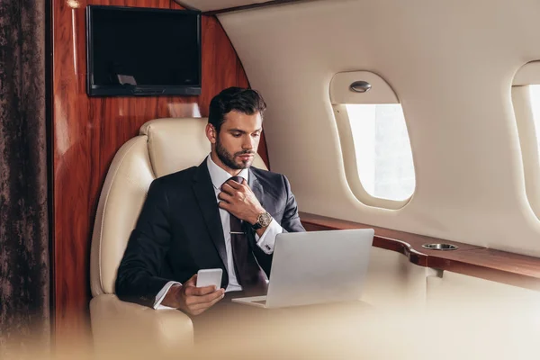 Selective focus of businessman in suit using laptop and holding smartphone in private plane — Stock Photo