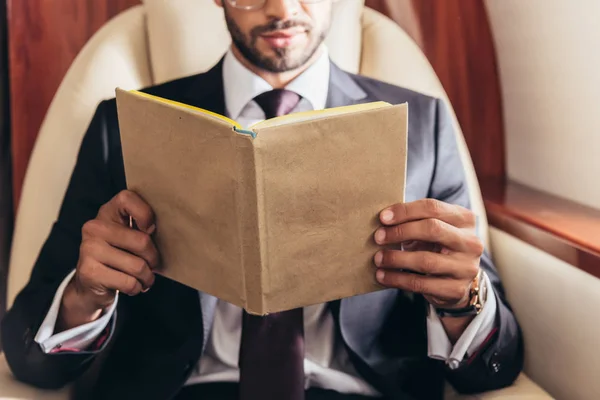 Ausgeschnittene Ansicht eines Geschäftsmannes im Anzug, der im Privatflugzeug Buch liest — Stockfoto