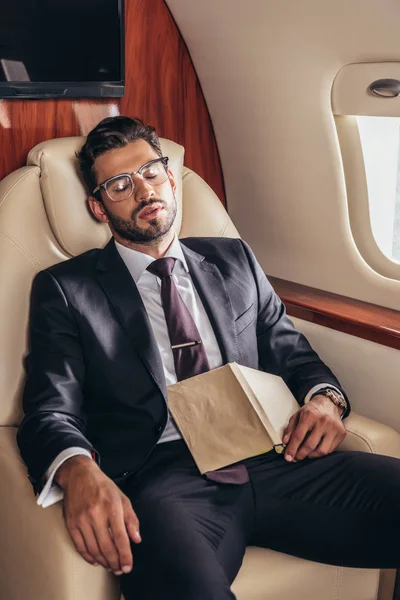 Bel homme d'affaires en costume avec livre dormant dans un avion privé — Photo de stock