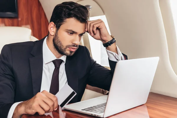 Handsome businessman in suit holding credit card and using laptop in private plane — Stock Photo