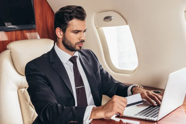 Красивий бізнесмен у костюмі тримає кредитну картку і використовує ноутбук у приватному літаку — стокове фото