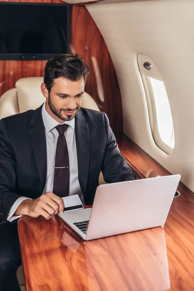 Schöner Geschäftsmann im Anzug mit Kreditkarte und Laptop im Privatflugzeug — Stockfoto