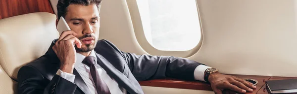 Panoramic shot of handsome businessman in suit talking on smartphone in private plane — Stock Photo