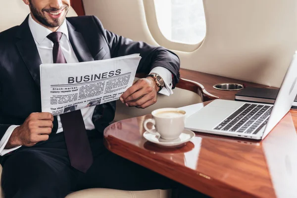 Cropped view of smiling businessman in suit reading business newspaper in private plane — Stock Photo