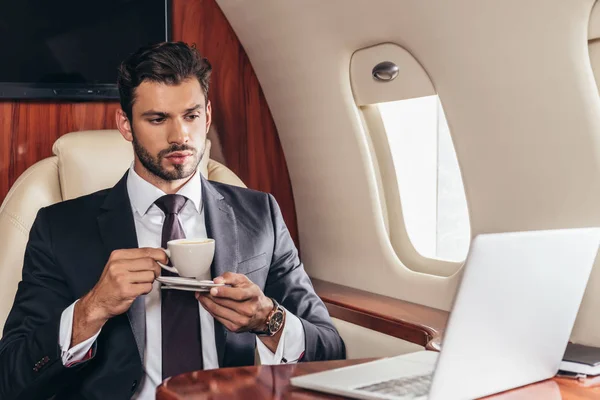 Guapo hombre de negocios en traje sosteniendo taza de café en avión privado - foto de stock