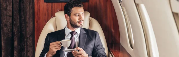 Panoramic shot of handsome businessman in suit holding cup of coffee in private plane — Stock Photo