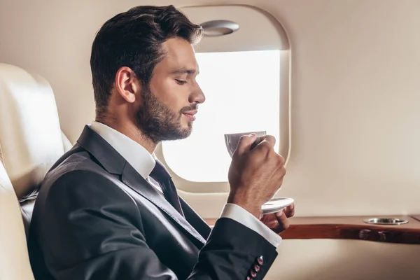 Vista lateral de empresário bonito em terno segurando xícara de café em avião privado — Fotografia de Stock