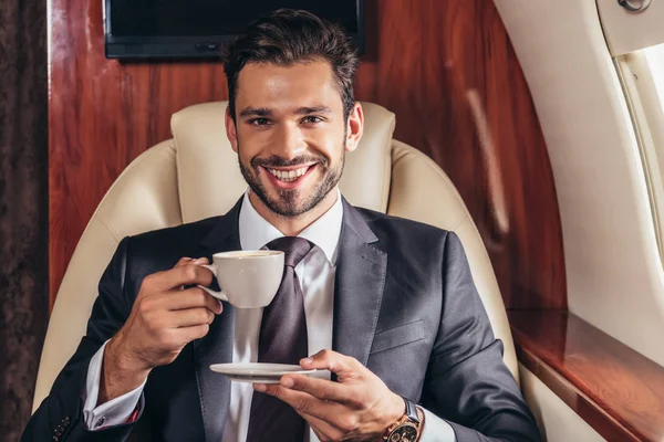 Homem de negócios bonito em terno sorrindo e segurando xícara de café em avião privado — Fotografia de Stock