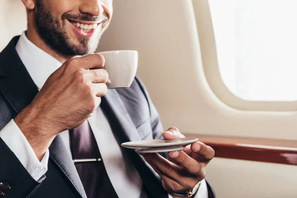 Vista ritagliata di un uomo d'affari sorridente in giacca e cravatta con una tazza di caffè in aereo privato — Foto stock