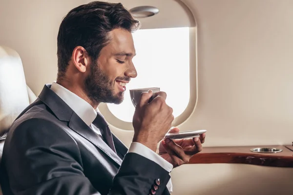 Vista laterale di un uomo d'affari sorridente in giacca e cravatta con una tazza di caffè in aereo privato — Foto stock