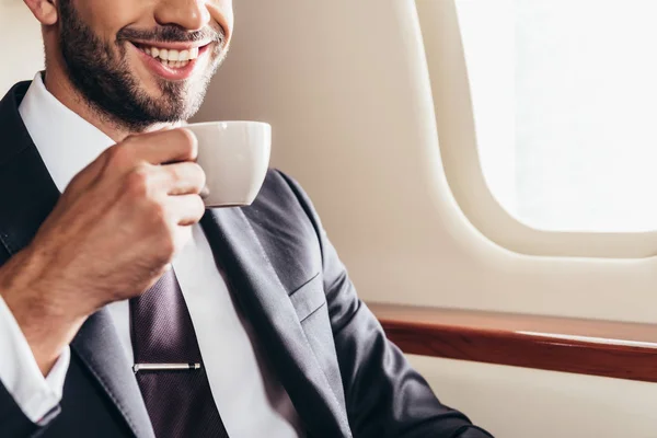 Vista ritagliata di un uomo d'affari sorridente in giacca e cravatta con una tazza di caffè in aereo privato — Foto stock
