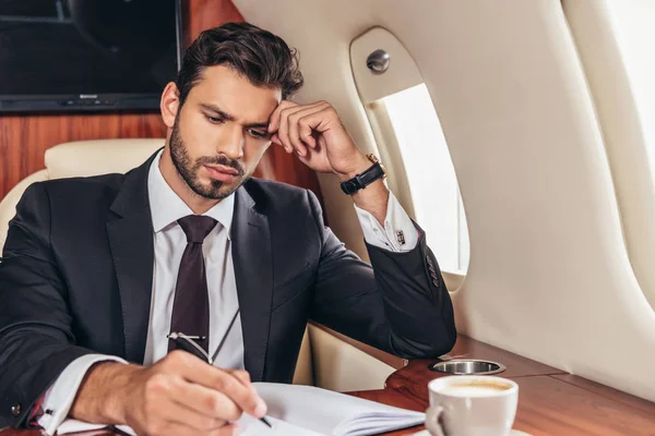 Guapo hombre de negocios en traje escrito en cuaderno en avión privado - foto de stock