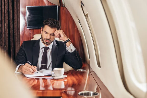 Красивый бизнесмен в костюме пишет в блокноте в частном самолете — стоковое фото