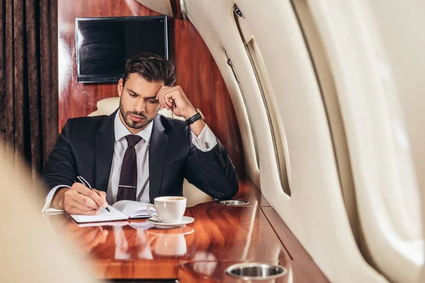 Guapo hombre de negocios en traje escrito en cuaderno en avión privado - foto de stock