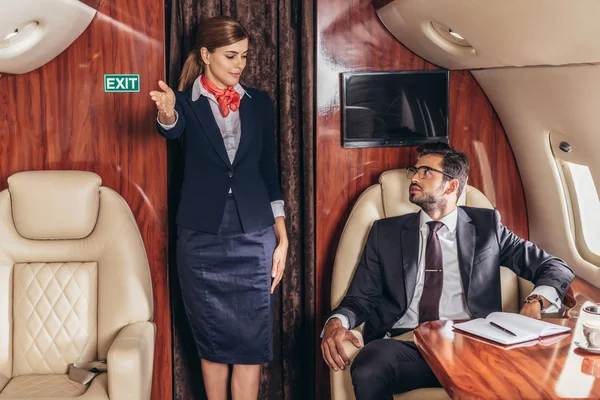 Asistente de vuelo mostrando gestos a guapo hombre de negocios en traje en avión privado - foto de stock