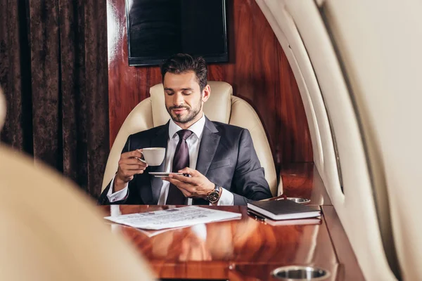Selective focus of handsome businessman in suit drinking coffee in private plane — Stock Photo