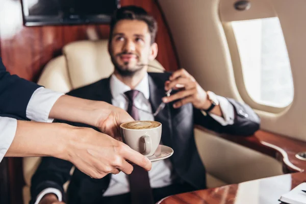 Vista ritagliata di assistente di volo dando tazza di caffè a bell'uomo d'affari in giacca e cravatta in aereo privato — Foto stock