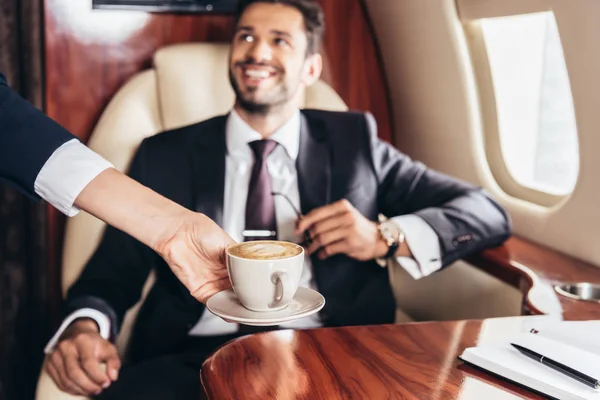 Vista cortada de comissário de bordo dando xícara de café para empresário bonito em terno em avião privado — Fotografia de Stock