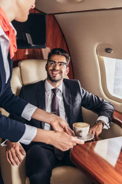Обрізаний вид бортпровідника, що дає чашку кави красивому бізнесмену в костюмі в приватному літаку — стокове фото