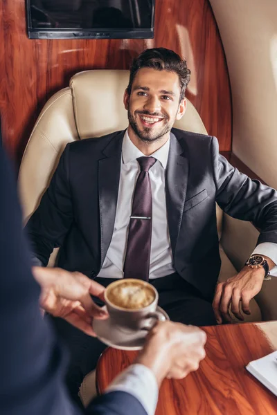 Vista cortada de comissário de bordo dando xícara de café para empresário bonito em terno em avião privado — Fotografia de Stock