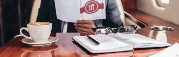 Panoramic shot of cup of coffee, notebook and glasses in private plane — Stock Photo