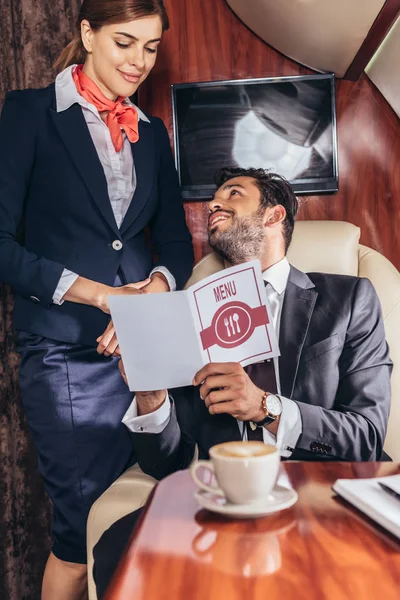 Красивый бизнесмен в костюме держит меню и разговаривает со стюардессой в частном самолете — стоковое фото