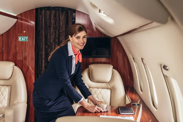 Asistente de vuelo sonriente poniendo taza de café en la mesa en avión privado - foto de stock