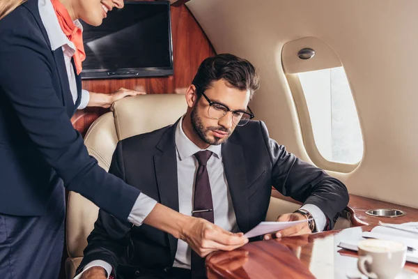 Vista cortada de comissário de bordo dando menu para empresário bonito em terno em avião privado — Fotografia de Stock