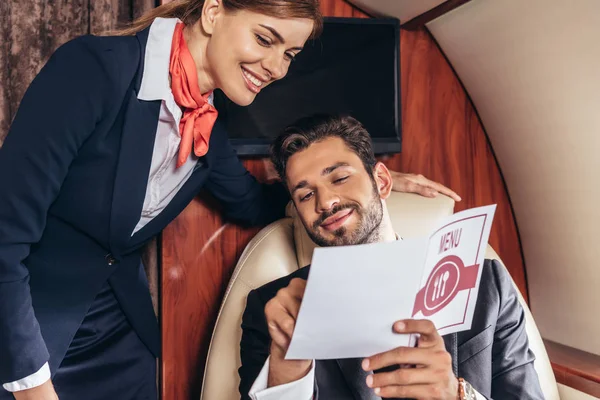 Bonito homem de negócios e comissário de bordo em uniforme olhando para o menu em avião particular — Fotografia de Stock