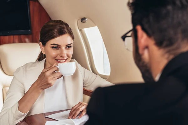 Sonriente empresaria bebiendo café y mirando a hombre de negocios hombre de negocios en avión privado - foto de stock