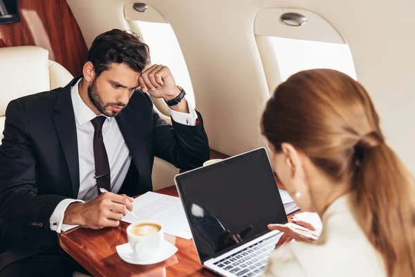 Бізнесмен дивиться на папір і ділову жінку, що використовує ноутбук у приватному літаку. — стокове фото