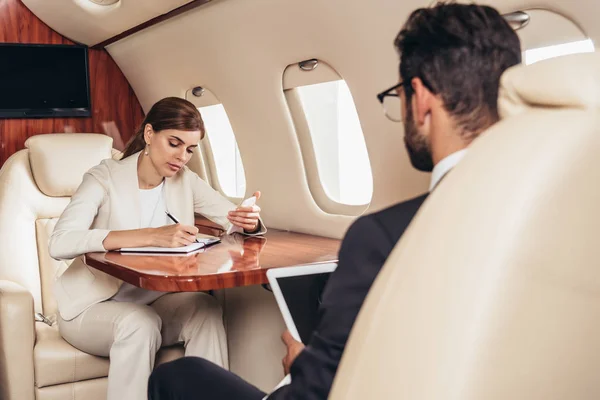 Hombre de negocios con tableta digital y empresaria escribiendo en cuaderno en avión privado — Stock Photo