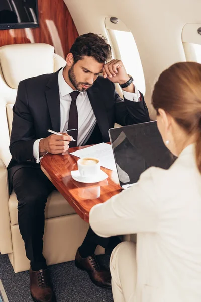 Бізнесмен дивиться на папір і бізнес-леді, використовуючи ноутбук в приватному літаку — стокове фото