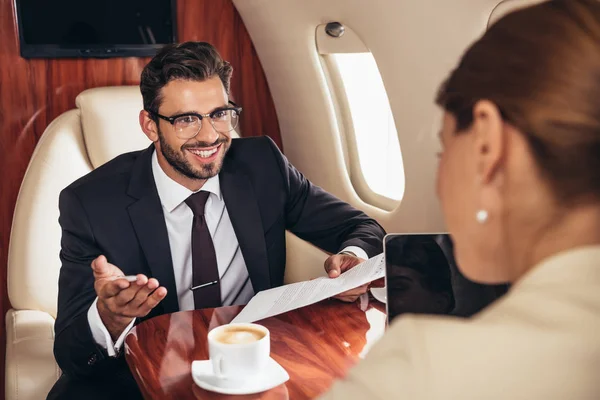 Selektiver Fokus eines lächelnden Geschäftsmannes, der Geschäftsfrau anschaut und Papier im Privatflugzeug hält — Stockfoto