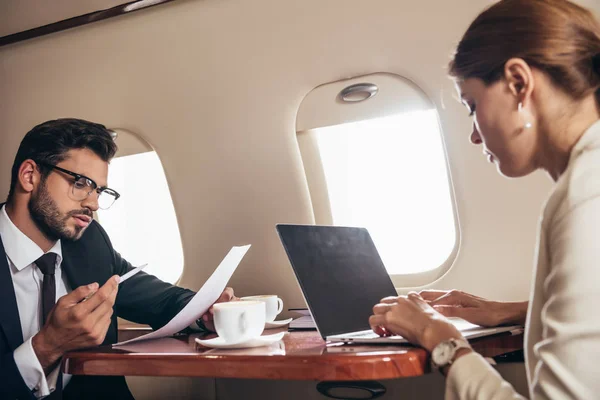 Hombre de negocios mirando documento y mujer de negocios utilizando el ordenador portátil en avión privado - foto de stock