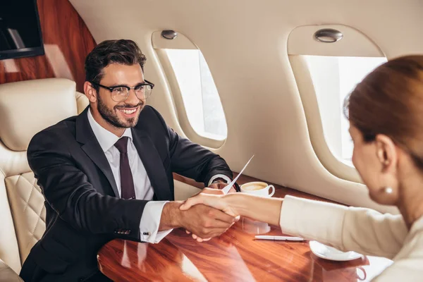 Sonriente hombre de negocios y mujer de negocios estrechando la mano en avión privado - foto de stock
