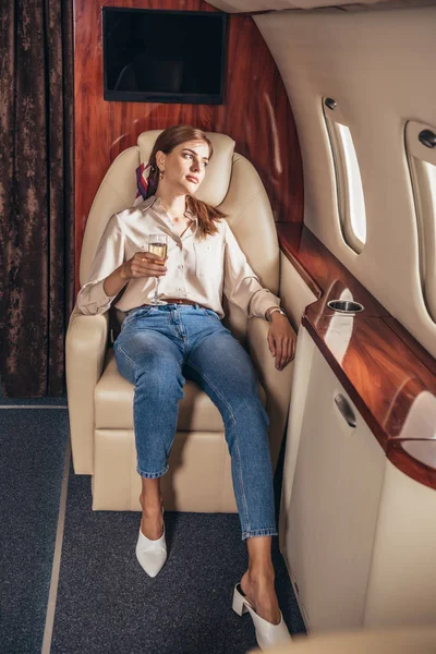 Attractive woman in shirt holding champagne glass and looking through window in private plane — Stock Photo