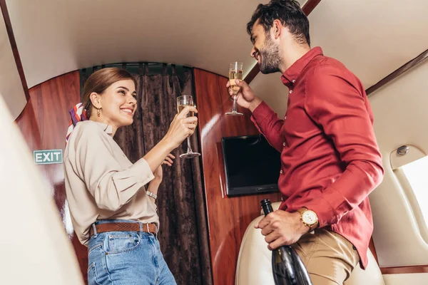 Улыбающийся парень с бутылкой и девушкой, держащий бокалы шампанского в частном самолете — стоковое фото