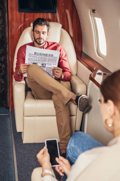 Foyer sélectif de petit ami lecture journal et petite amie tenant smartphone dans l'avion privé — Photo de stock