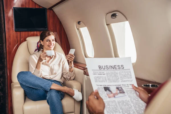 Selektiver Fokus des Freundes, der Zeitung liest und Freundin, die Tasse und Smartphone im Privatflugzeug hält — Stockfoto