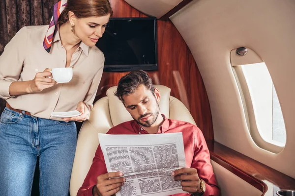 Novio y novia con taza de leer periódico en avión privado - foto de stock