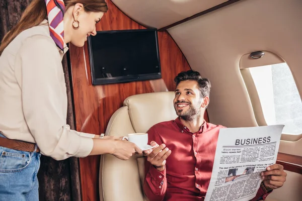 Fidanzata dando tazza di caffè al fidanzato con gli affari di giornale in aereo privato — Foto stock